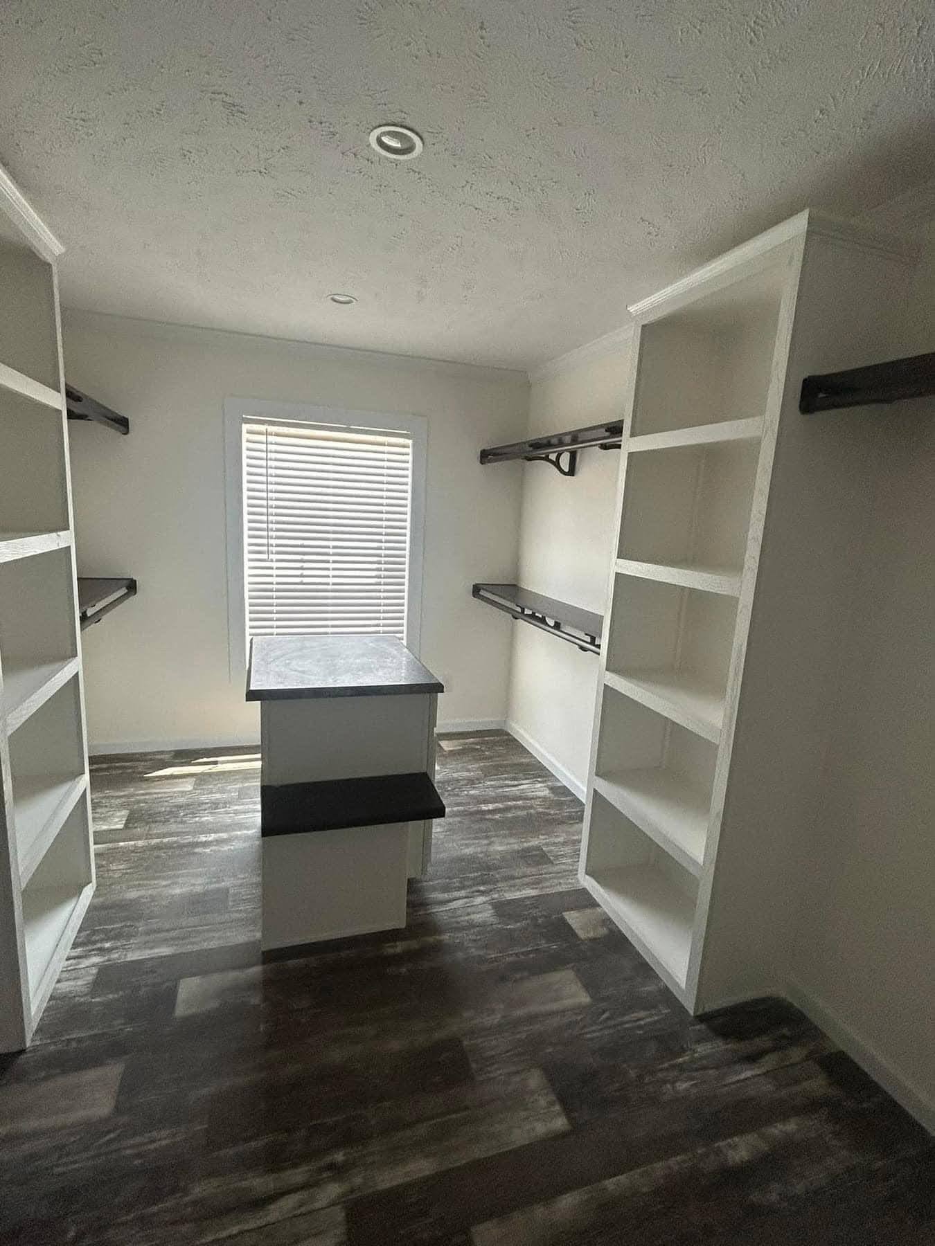 A spacious walk-in closet with dark wooden floor, white shelves, and rods. Natural light filters through a window with blinds, creating a cozy ambiance.