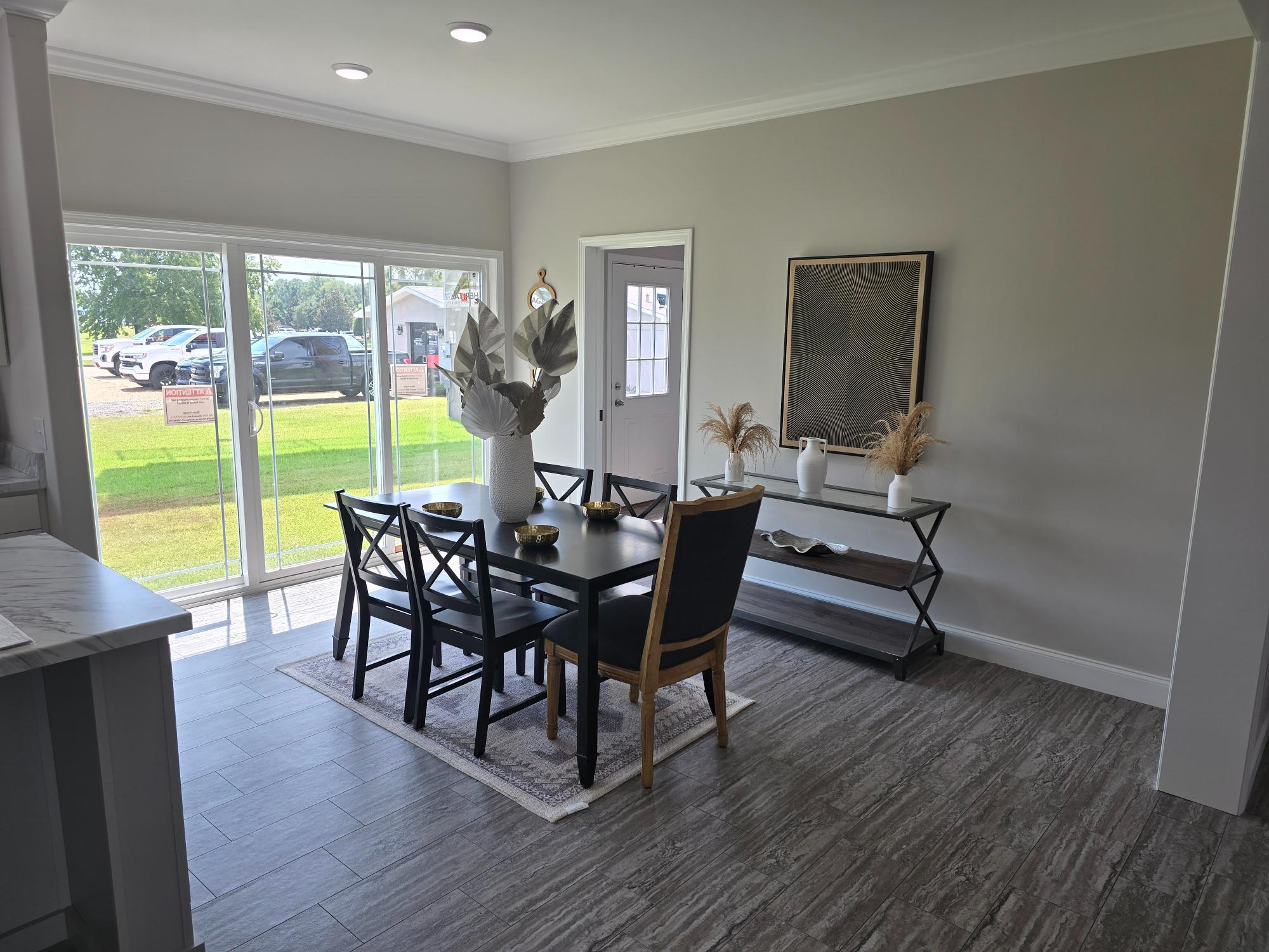 Modern dining room with a black table, six chairs, and a white vase with foliage. Large windows offer views of a green yard; bright and airy ambiance.