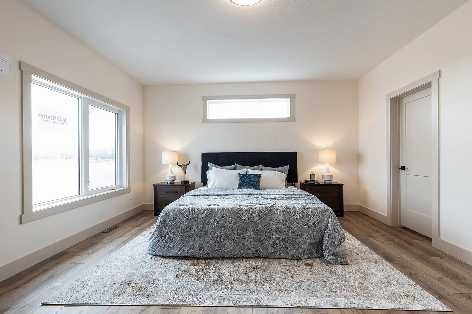 A cozy bedroom with a large bed featuring patterned gray bedding and multiple pillows. Two bedside tables with lamps create a warm, inviting tone.