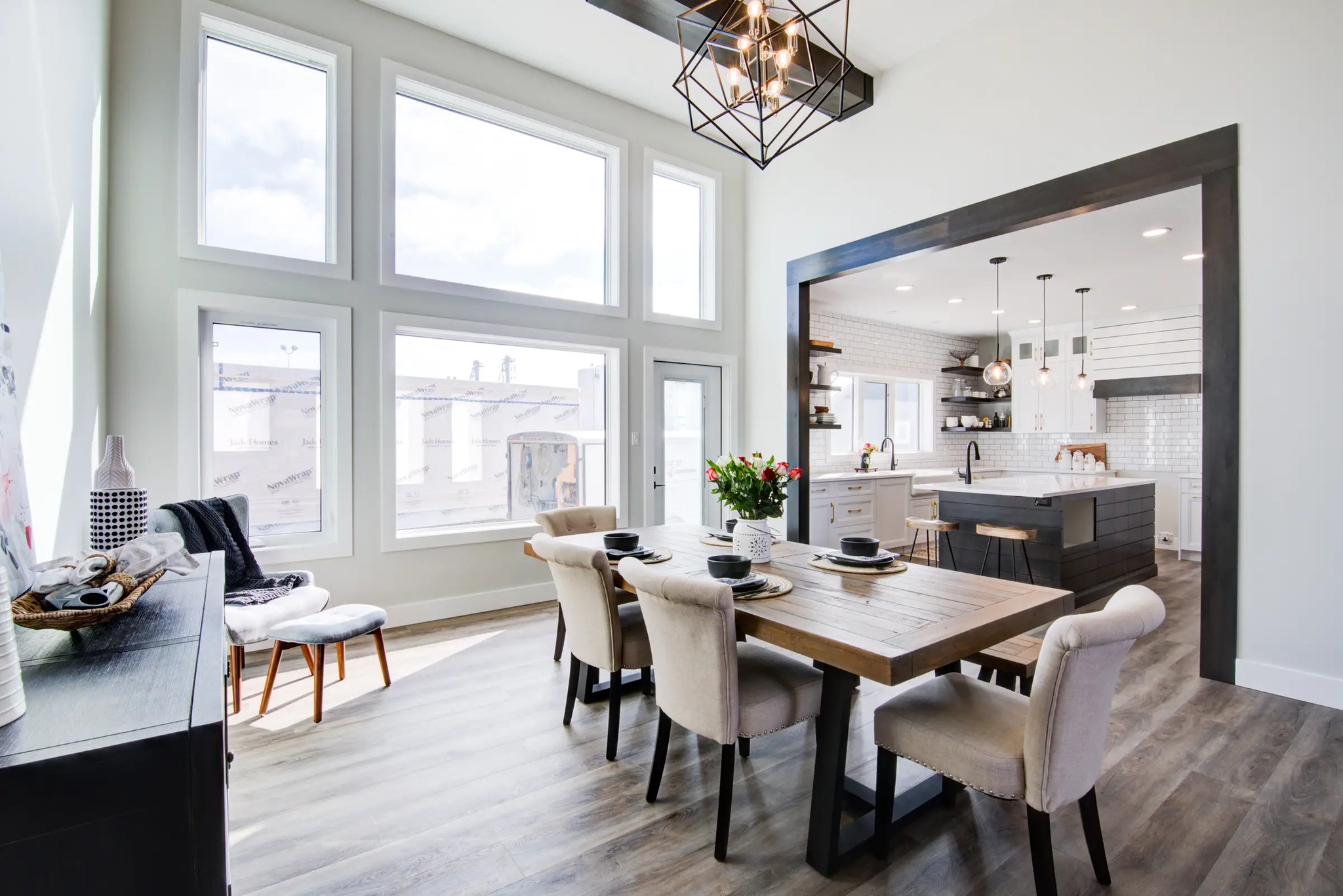 Spacious, bright dining area with a rustic wood table, beige chairs, and large windows. Kitchen in the background, modern decor with pendant lights.