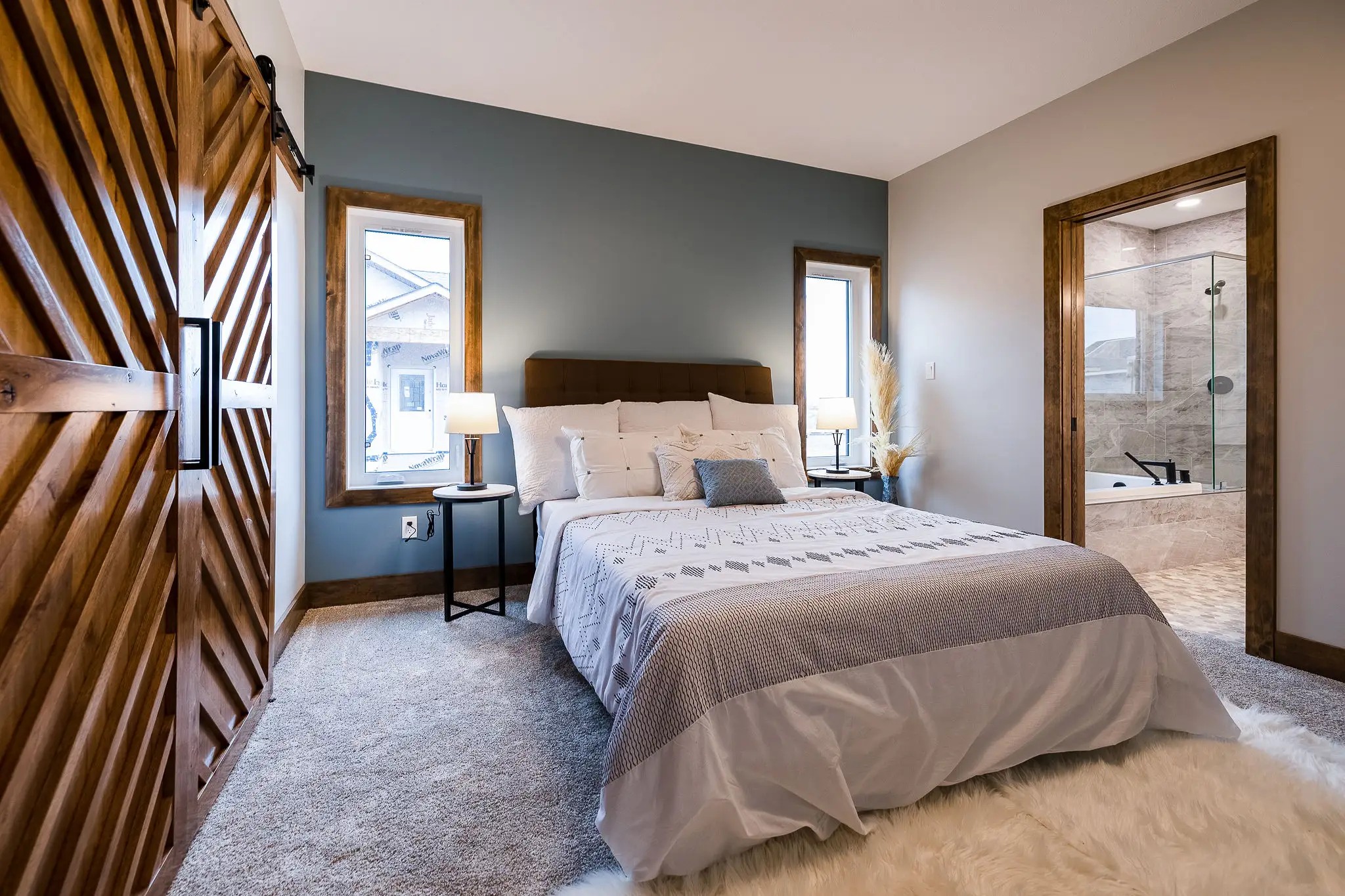 A cozy bedroom with a modern style features a bed with white and gray bedding, flanked by two windows. A sliding wooden barn door leads to a bathroom. The atmosphere is serene and inviting.