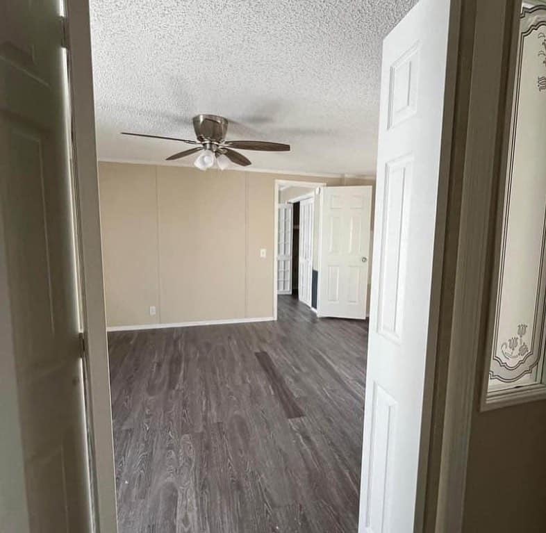 Empty room with beige walls and dark wood flooring. A ceiling fan with light hangs in the center, and three open white doors lead to other rooms.