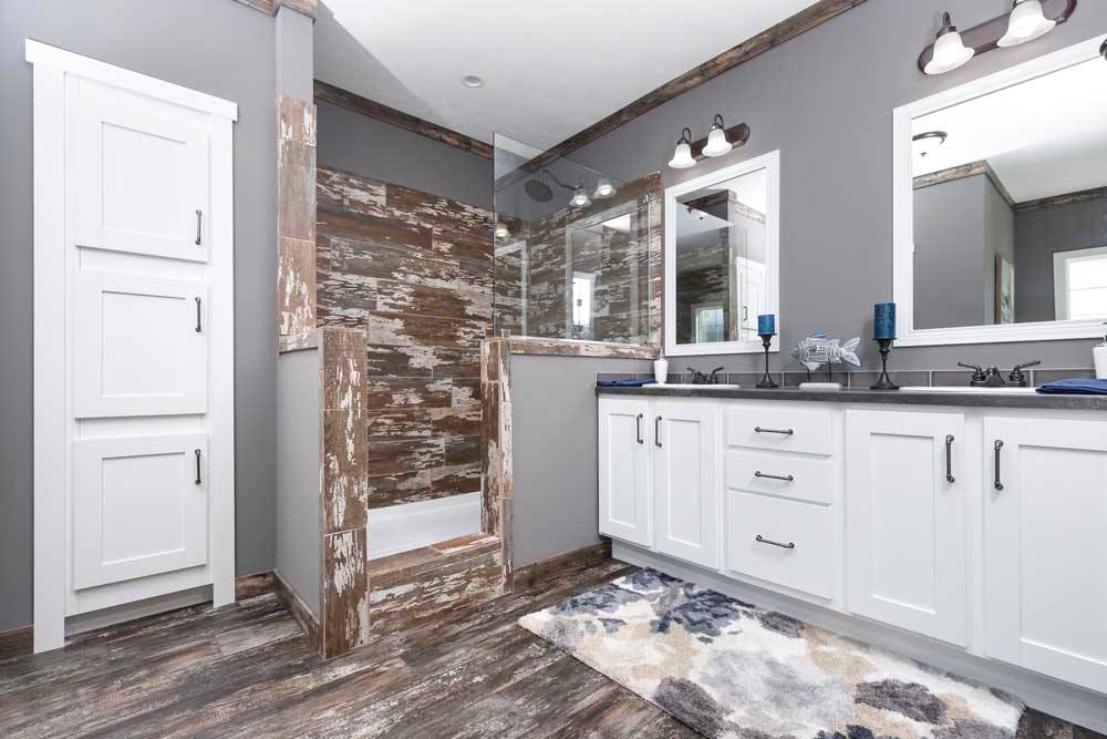 Modern bathroom with gray walls, featuring a rustic wooden-tiled shower, white cabinets, dual sinks with mirrors, and a patterned rug on wood flooring.