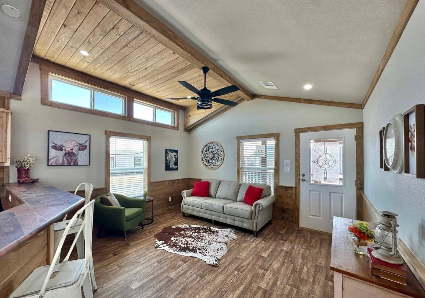 Cozy living room with a high wood ceiling, ceiling fan, and large windows. Features a gray couch with red pillows, green chair, cowhide rug, bar stools, and rustic decor.