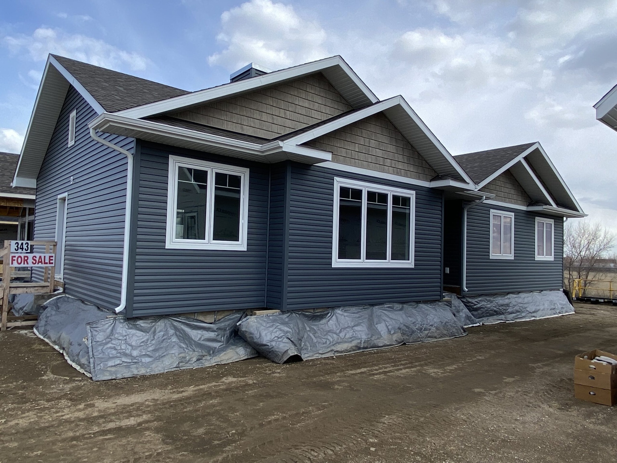 Gray house with white-trimmed windows and gabled roof, set on a dirt lot with tarps at the base. A "For Sale" sign is visible, under a cloudy sky.