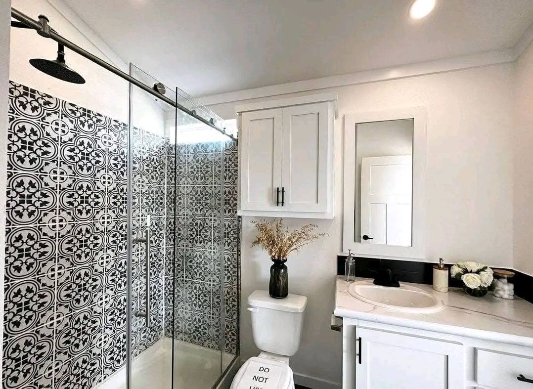 Modern bathroom with black and white patterned shower tiles, glass door, white cabinetry, and a vase with dried flowers. Bright, clean, stylish.