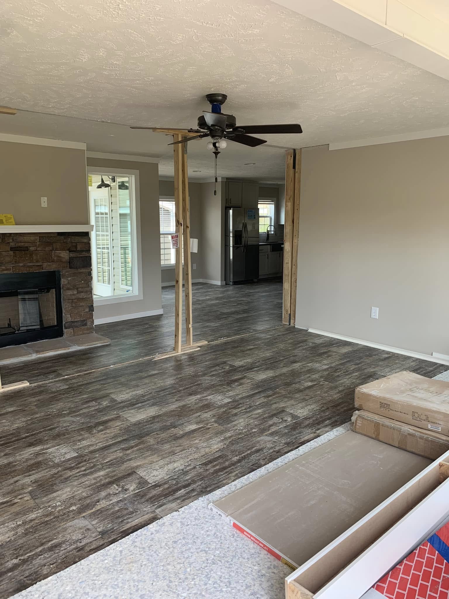 A partially renovated living room with dark wooden floors, ceiling fan, stone fireplace, and kitchen visible. Wooden supports are in place, suggesting ongoing construction.