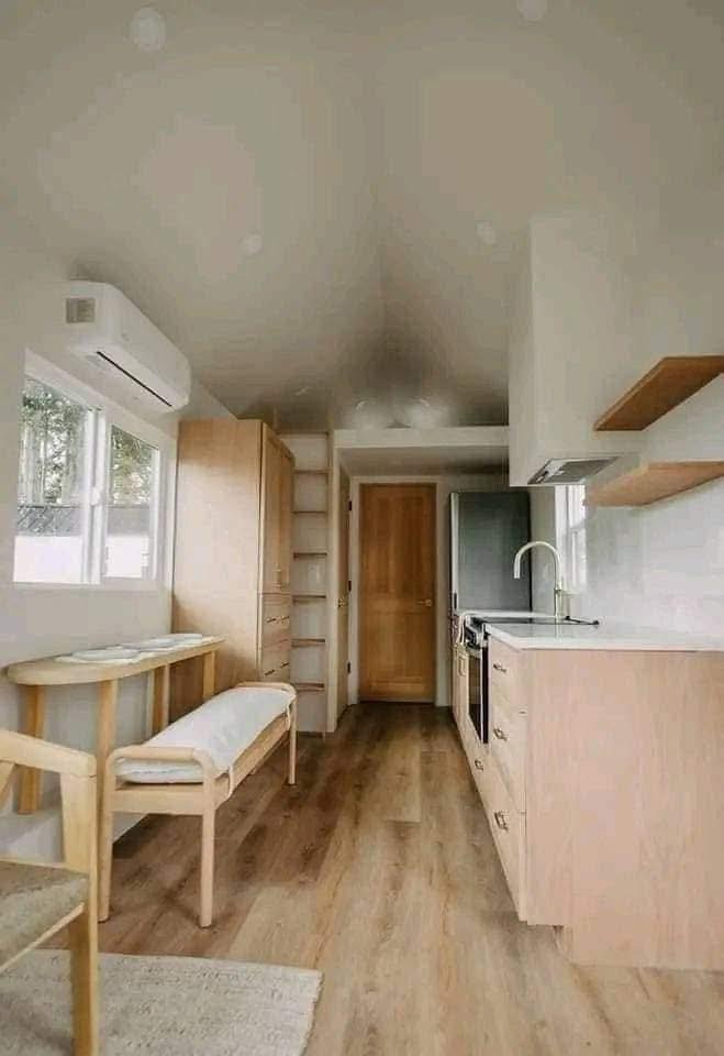 Minimalist tiny home kitchen with light wood accents. Features include wood floors, a compact dining area with bench seating, and ample natural light from windows. The space feels airy and organized.