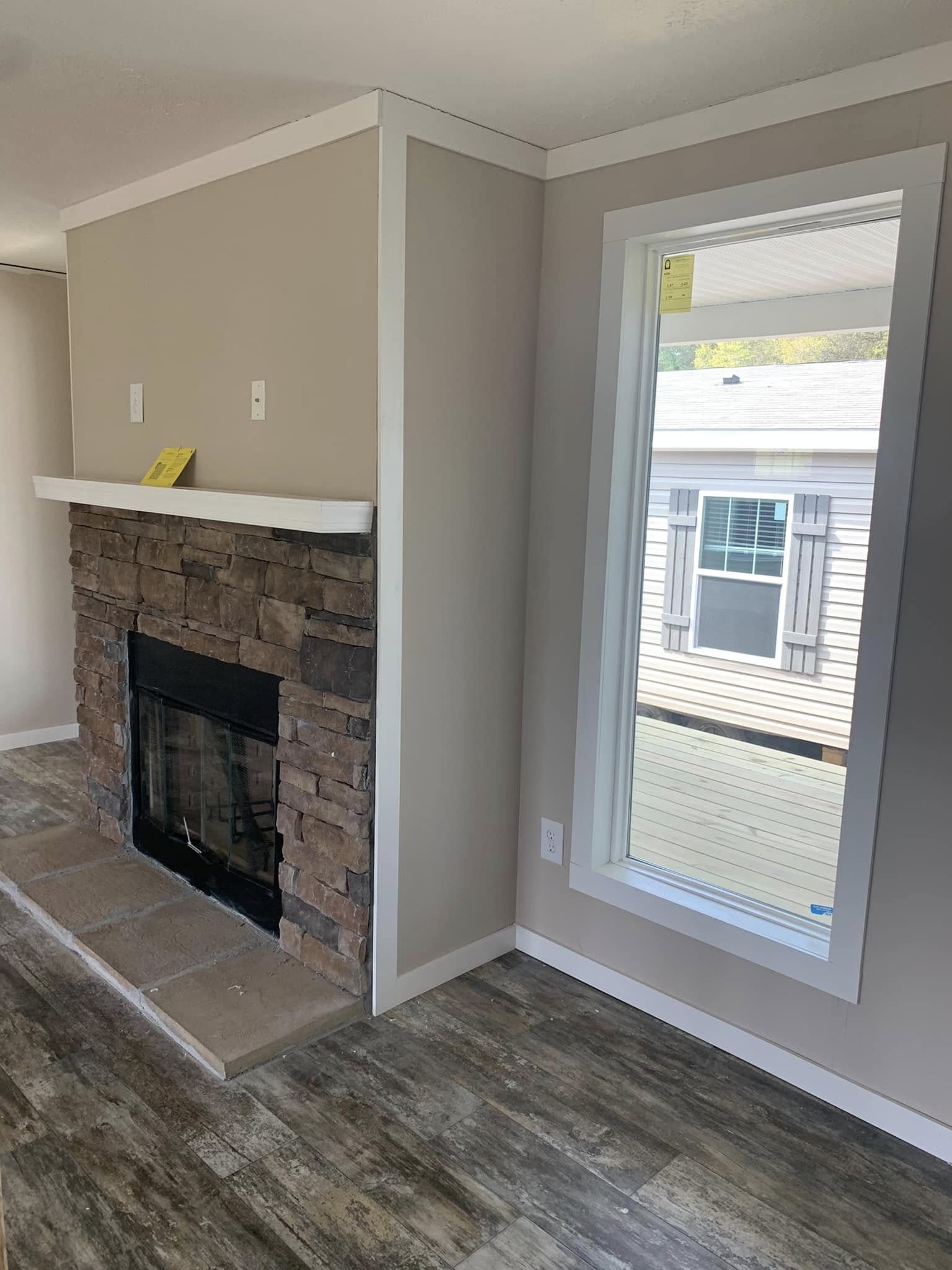 A cozy room features a stone fireplace with a wooden mantle on the left, and a large window on the right, allowing natural light to brighten the space.
