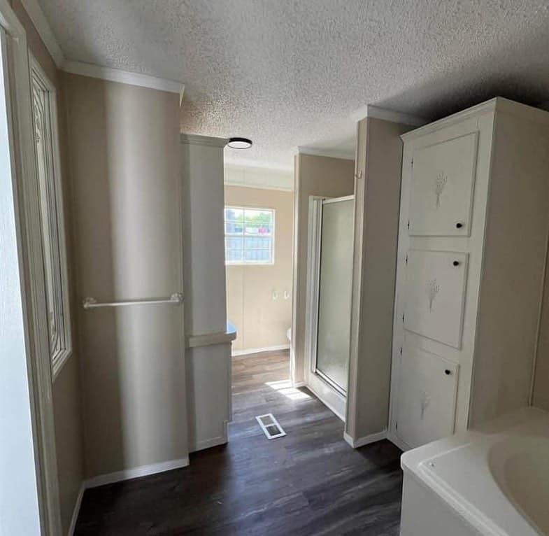 Narrow bathroom with dark wood floor, white fixtures, and beige walls. Features a shower, storage cabinet, and a window letting in natural light.