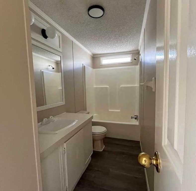 Compact bathroom with white walls and fixtures. Features a vanity sink, a mirror with lights, a small window, bathtub, and wooden floor. Minimalist design.