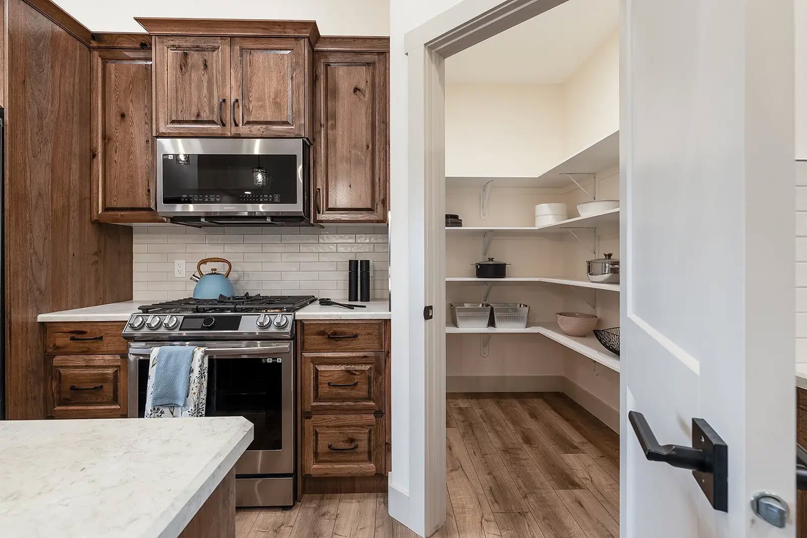 Cozy kitchen with wooden cabinets, stainless steel stove, and a blue kettle. Open door reveals a pantry with white shelves and various kitchenware.