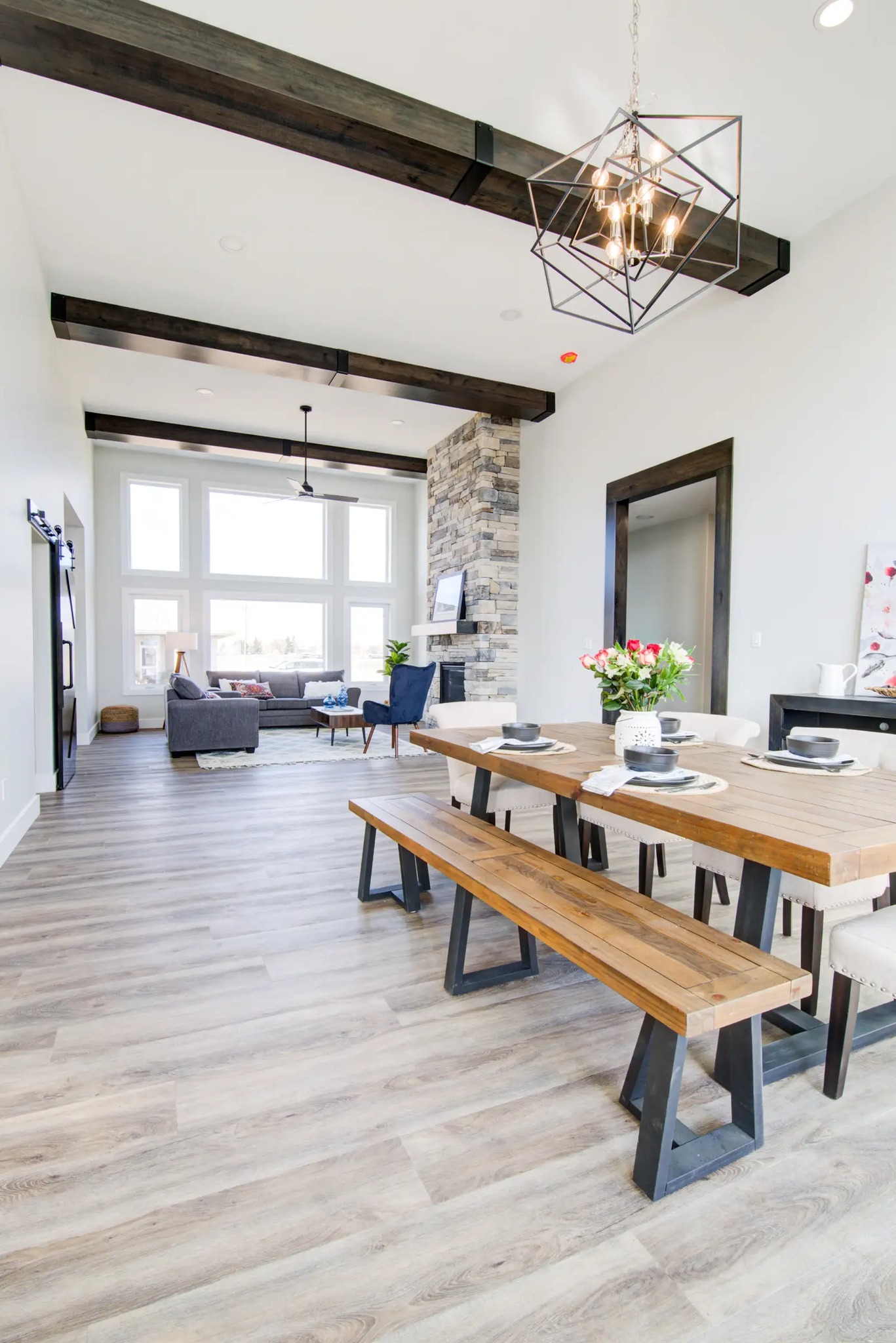 Modern open-plan living room with high ceilings and wooden beams. Features a rustic dining table, stone fireplace, large windows, and cozy seating.