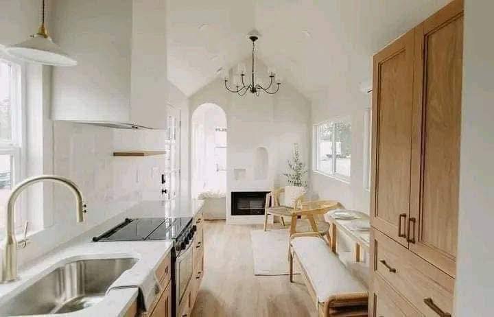 Bright, minimalist living space with light wood furniture, a chandelier, and arched window. Cozy seating area by a small fireplace. Warm and inviting.