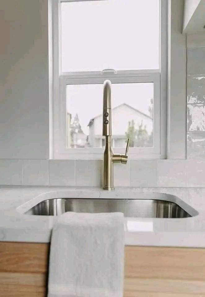 Modern kitchen sink with a sleek, gold-tone faucet, set against a white countertop. A soft towel hangs over the edge, and a bright window provides natural light.