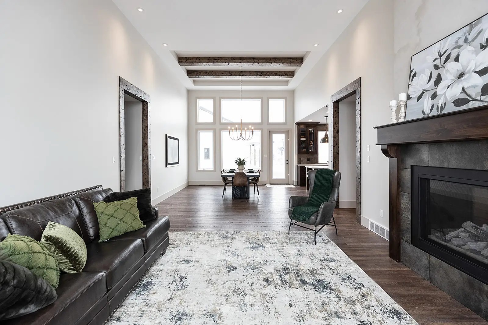 A modern living room with high ceilings, featuring a black leather sofa with green cushions on the left, a patterned gray rug, and a fireplace on the right. Large windows illuminate a dining table in the background. The room conveys a cozy and elegant atmosphere.