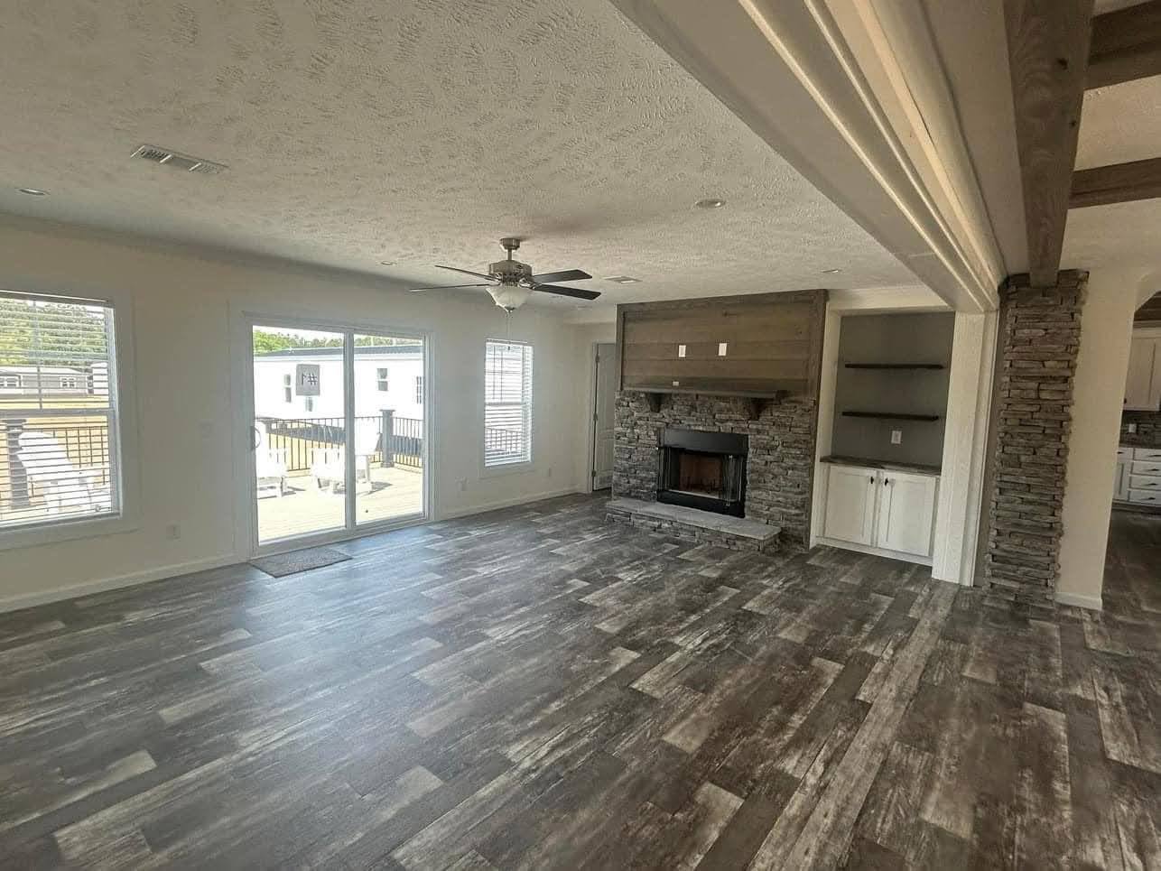 Spacious living room with rustic wood flooring, a stone fireplace, ceiling fan, and large windows leading to a sunny outdoor deck. Cozy and inviting.