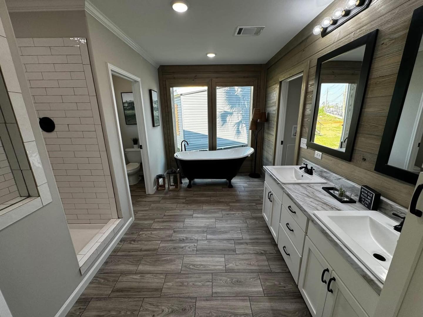 Modern bathroom with wood-look tile flooring, a freestanding black tub by a large window, dual sinks with mirrors, and a glass-door shower.