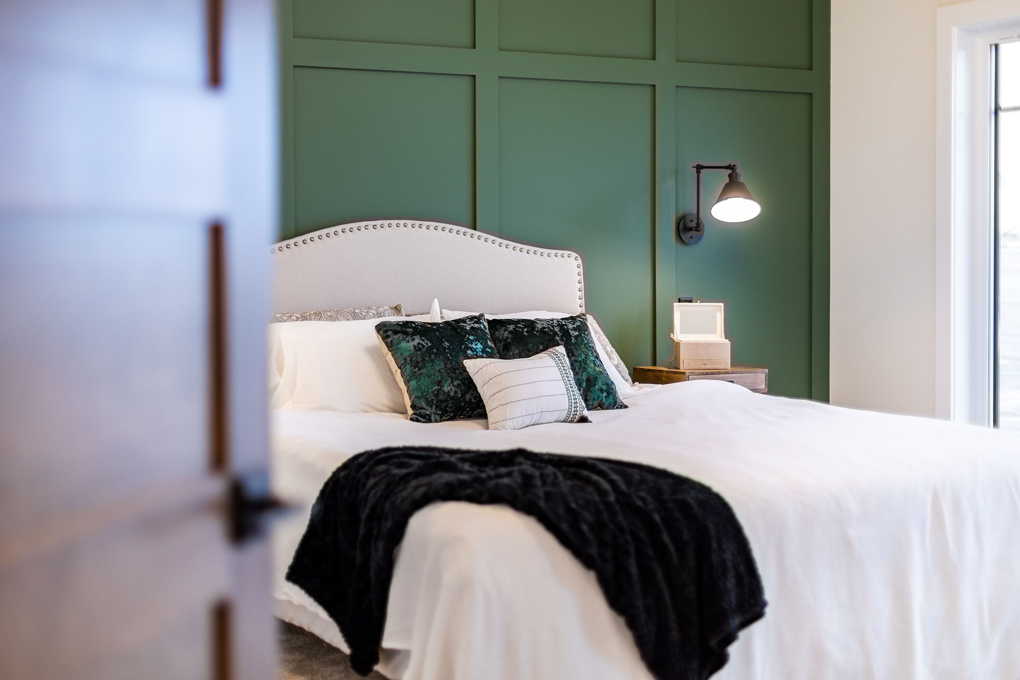 A cozy bedroom with a white bed, dark teal pillows, and a black throw. The wall is paneled green, with a wall-mounted lamp adding warmth.