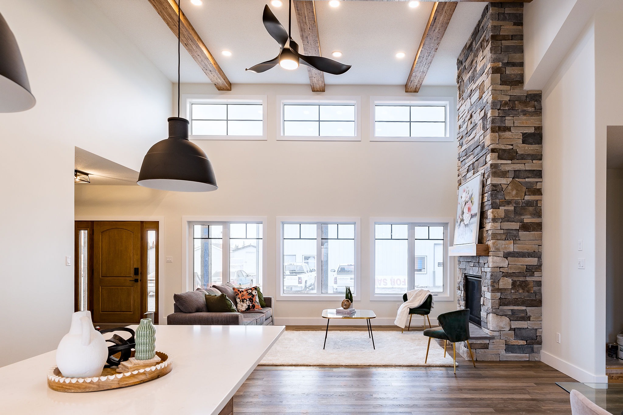 Modern living room with high ceilings, wood beams, and large windows. Features a stone fireplace, cozy seating, and elegant decor. Bright, airy atmosphere.
