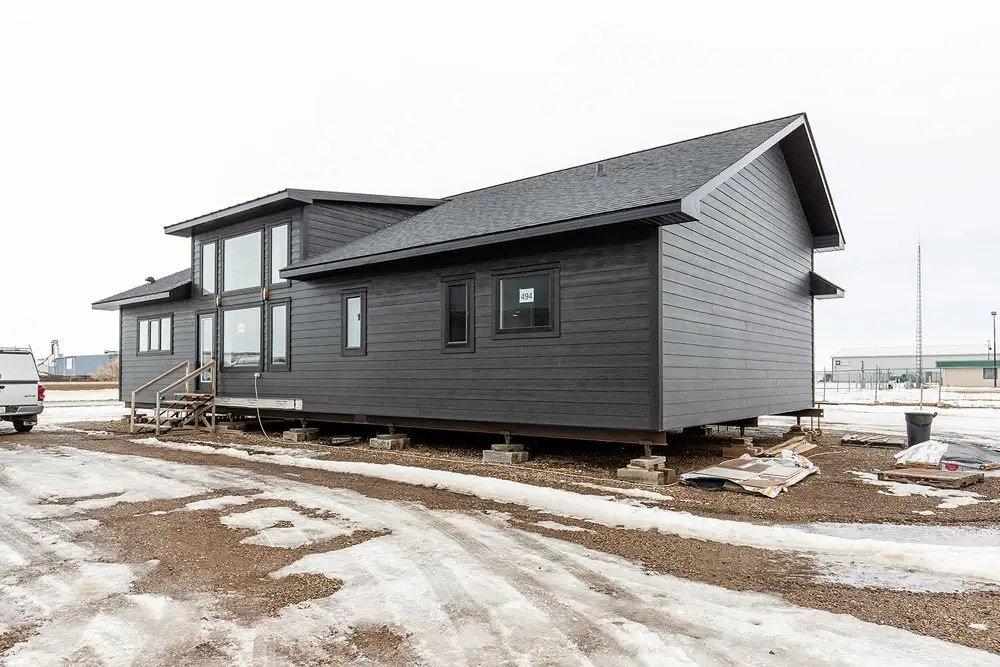 A modern, dark gray prefab home sits elevated on concrete blocks amidst a snowy, muddy landscape. A pickup truck is parked nearby, and the sky is overcast.