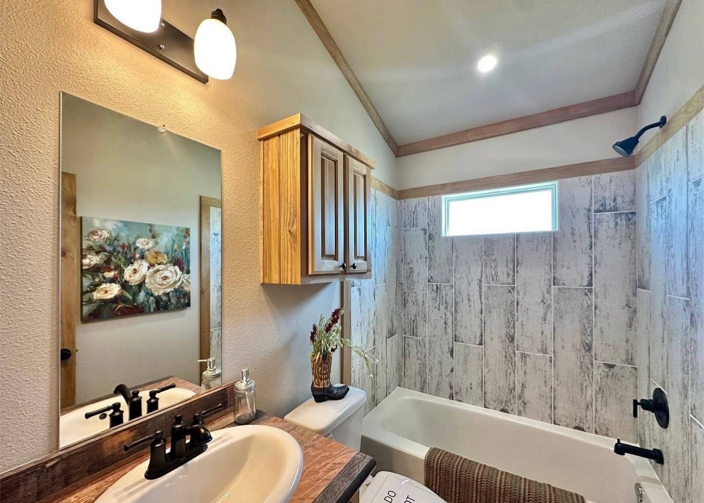 Cozy bathroom with wood accents, a white bathtub under a high window, and a rustic vanity. Ambient lighting and floral art add warmth.