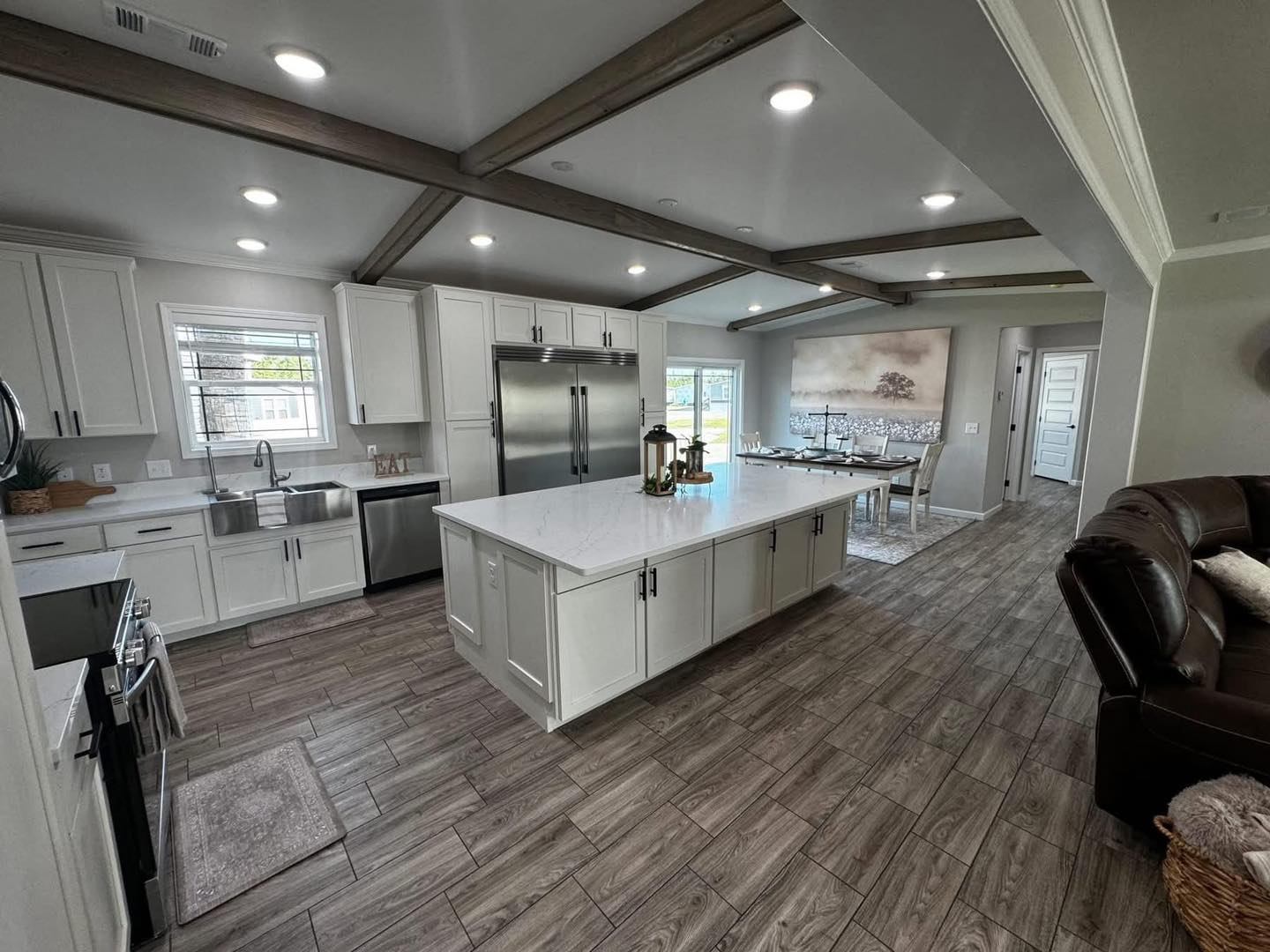 Spacious kitchen with a white island, stainless steel appliances, and rustic wood beams. Opens to a dining area with a large painting, creating a warm, inviting atmosphere.