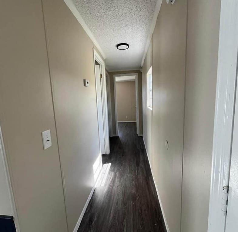 Narrow hallway with beige walls, dark wooden floor, and white ceiling. Natural light enters through a window, creating a calm, minimalist atmosphere.