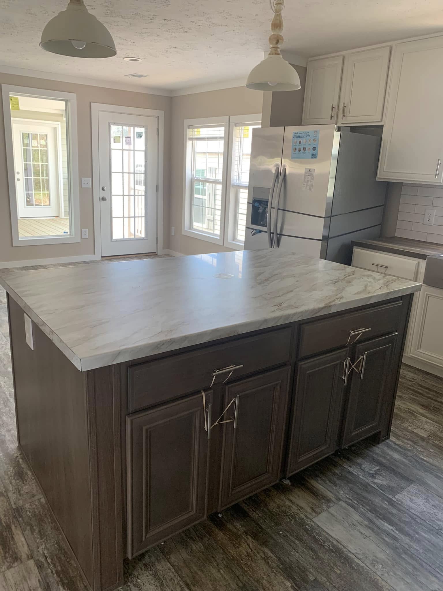 Bright kitchen with a large marble island, dark wood cabinets, and pendant lights. French doors and windows provide natural light, enhancing a modern, airy feel.