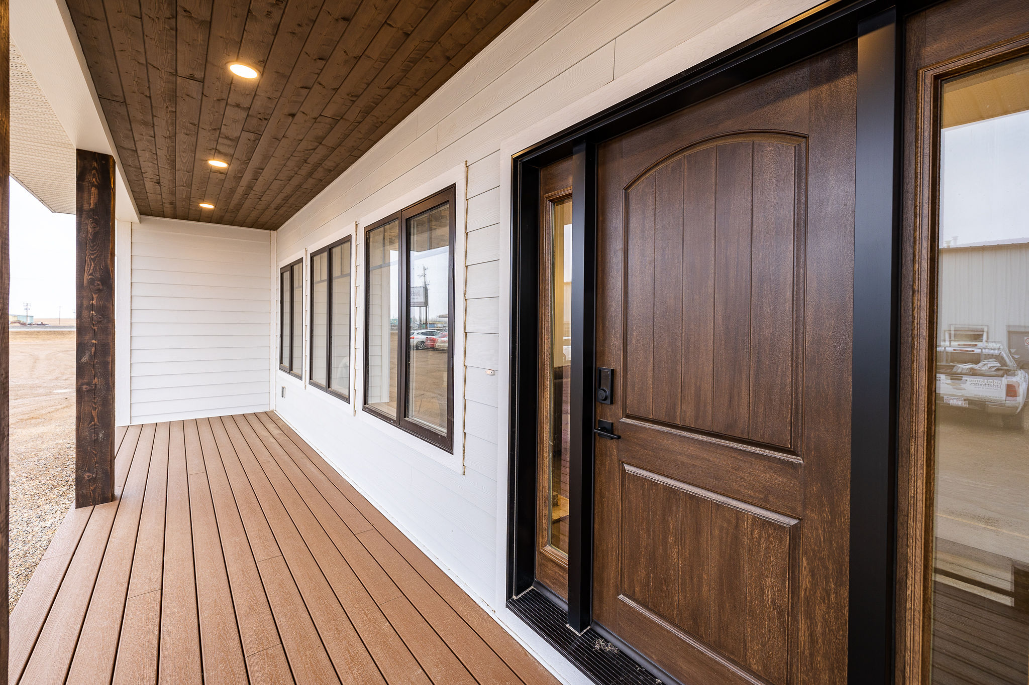 Covered porch with brown wooden decking and ceiling, white siding, rectangular windows lined up, and a large wooden front door. Modern and inviting.