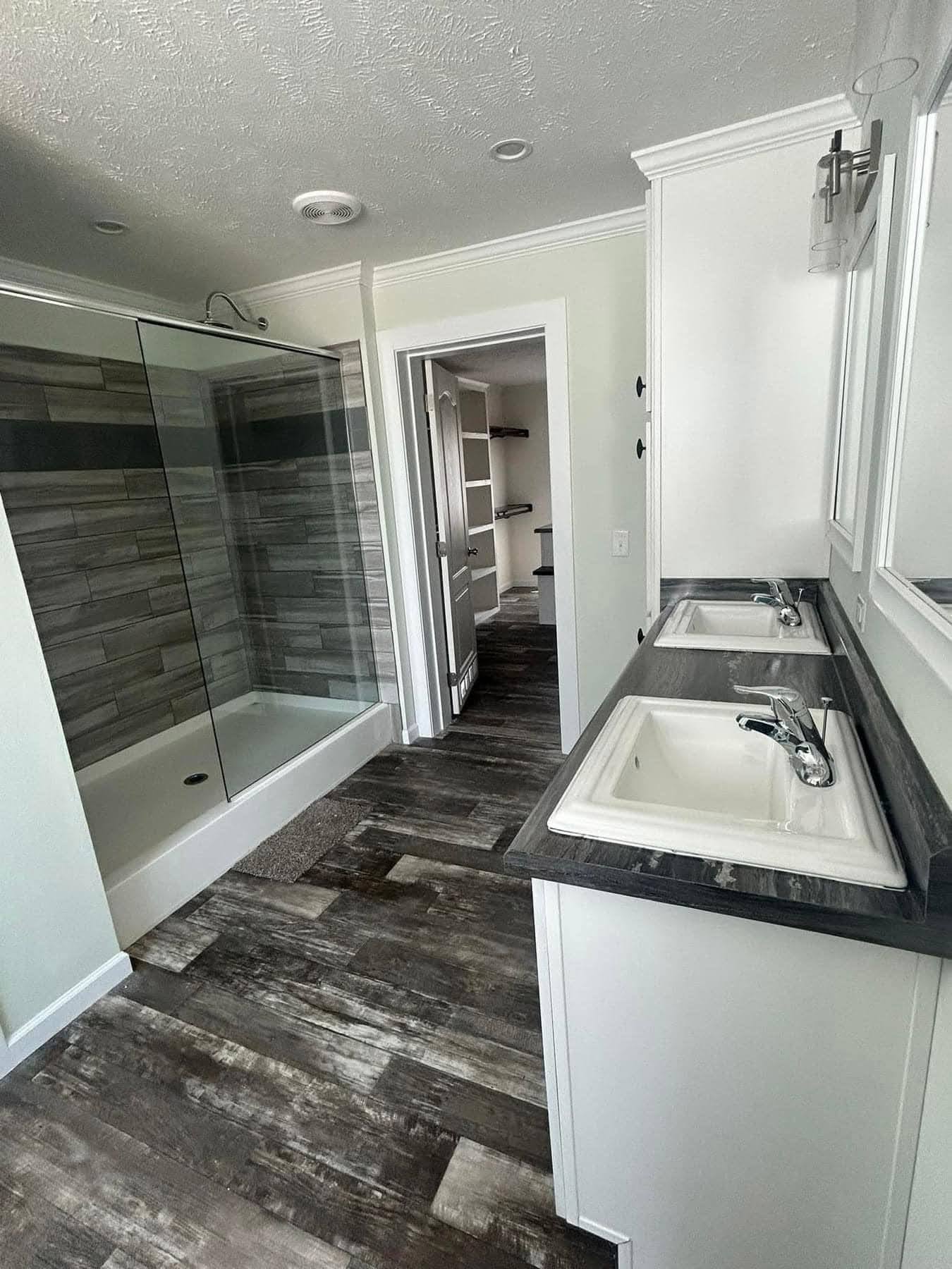 Modern bathroom with textured gray tiles in the shower, dual sinks on a dark counter, and rustic wood floor. Bright and spacious ambiance.