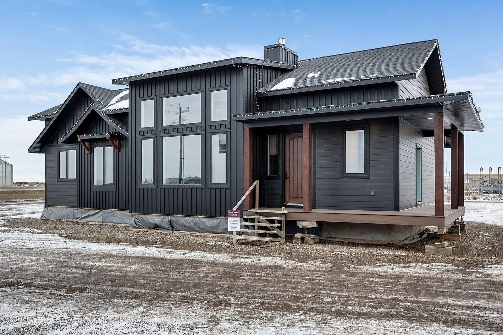 Modern, dark grey modular home on a snowy lot under a clear blue sky. Features large windows and a wooden porch, conveying a sleek, minimalist vibe.
