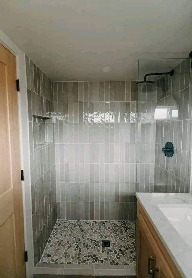 A modern shower with vertical gray tiles and a pebble stone floor. Features a glass door, black fixtures, and a natural wood vanity, creating a calm ambiance.