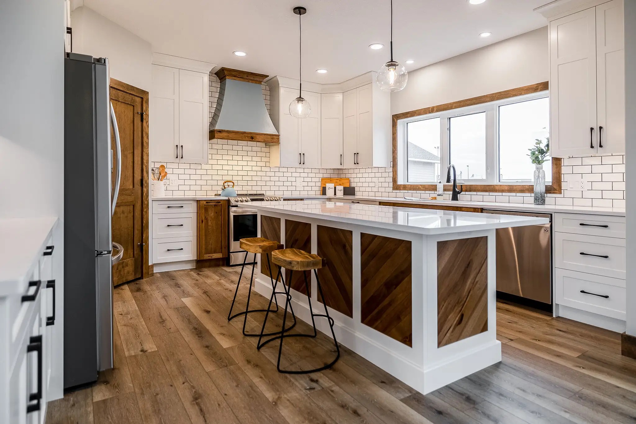 Modern kitchen with white cabinets, wood accents, and a large island with two stools. Subtle lighting, subway tile backsplash, and stainless steel appliances.