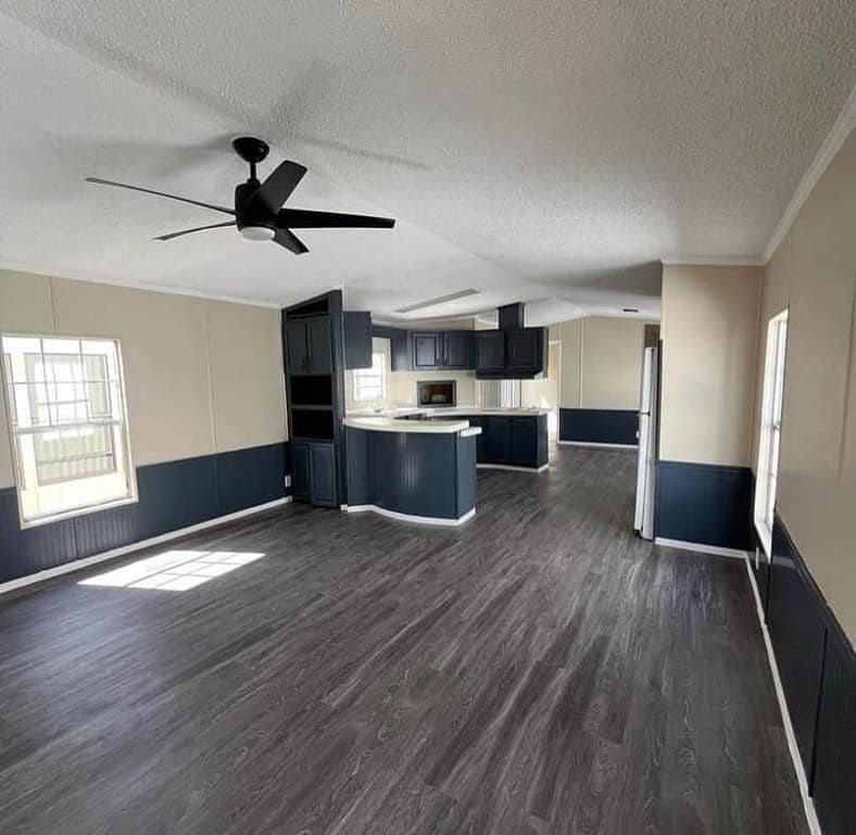 Open-concept living space with dark laminate flooring, beige walls, navy wainscoting, and a modern black ceiling fan. Kitchen with navy cabinets and white counters.