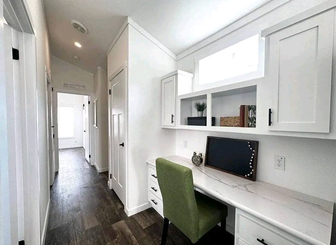 Bright hallway with white walls and cabinetry, featuring a marble desk and green chair. Shelves hold a plant and books. The tone is modern and clean.