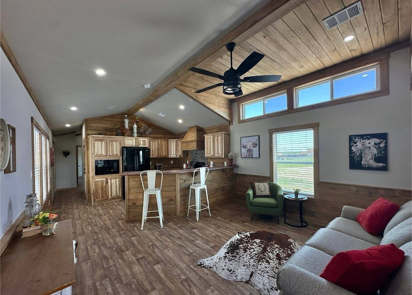 Spacious, rustic living room with wooden ceiling and floors, modern kitchen. Cozy sofa with red cushions, cowhide rug, green chair, and wall art.
