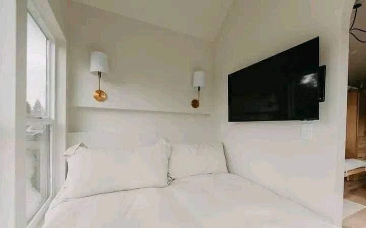 A cozy bedroom corner featuring a neatly made bed with fluffy white bedding. Two modern wall lamps with gold accents and a flat-screen TV add elegance.