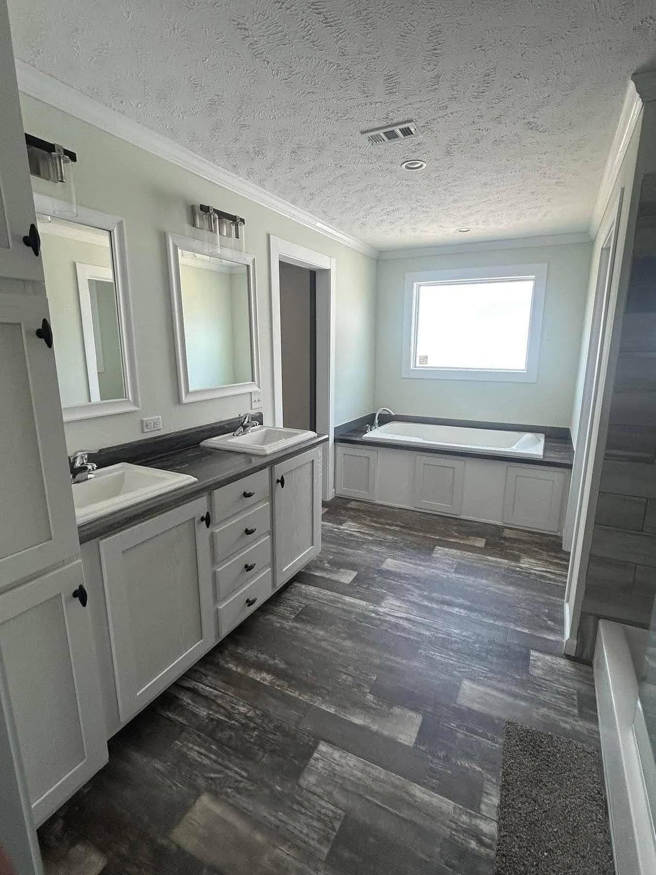 A spacious bathroom with two sinks and mirrors on the left. A large bathtub is beneath a window on the right, with wood-style flooring throughout.