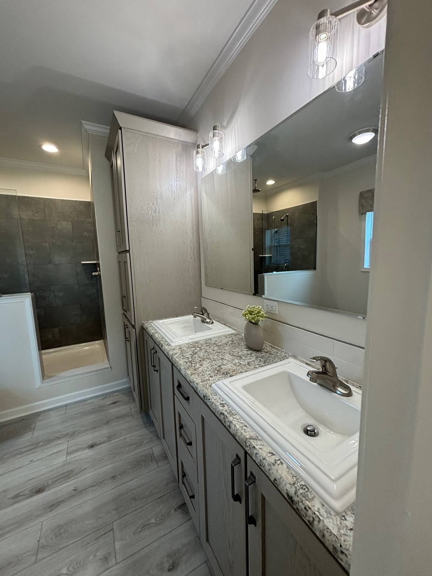 Modern bathroom with dual sinks on a speckled granite countertop, large mirror, light wood cabinets, gray tile shower, and elegant lighting.