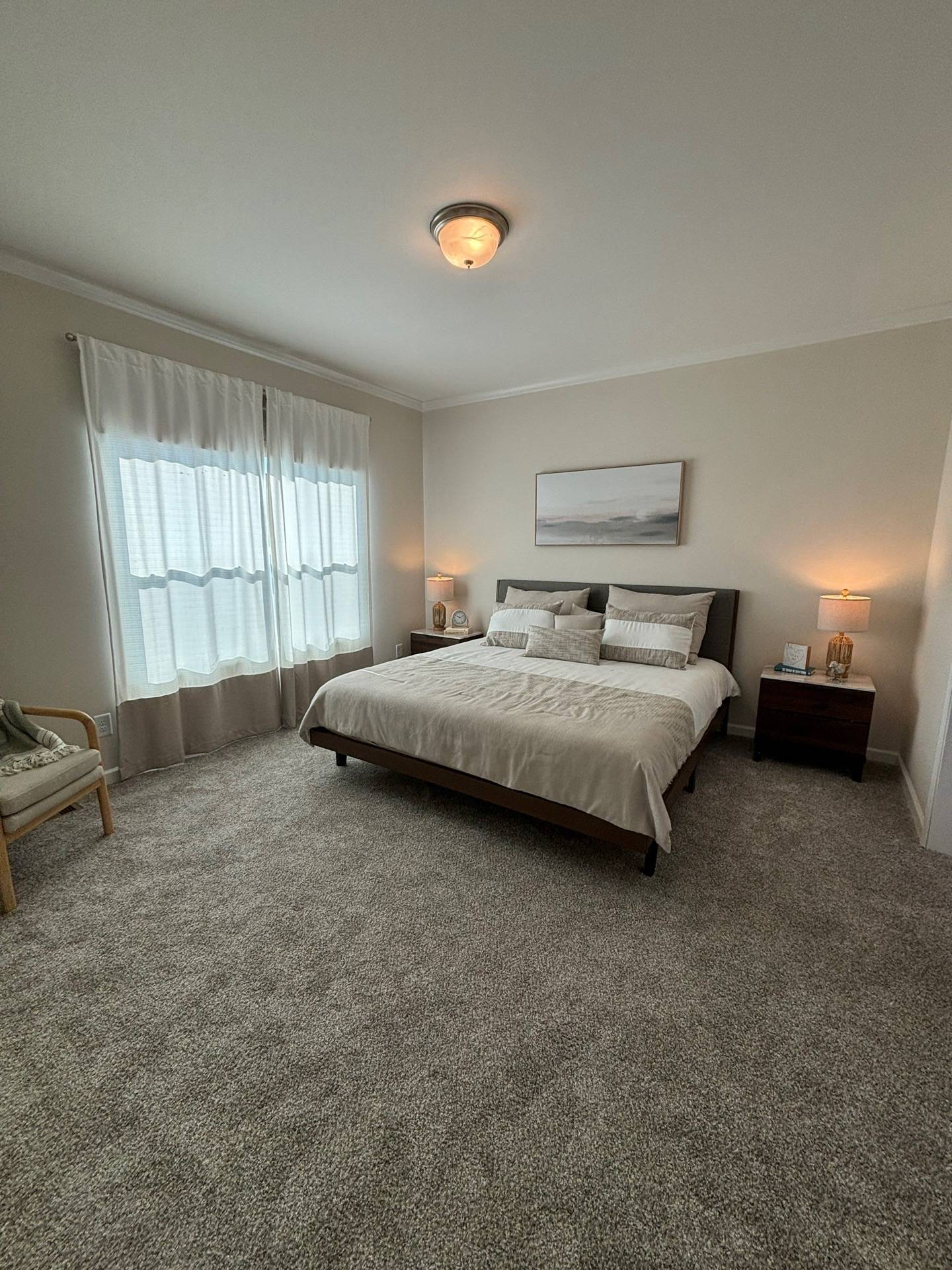 Spacious bedroom with neutral tones, featuring a neatly made king-size bed, two bedside tables with lamps, a large window with sheer curtains, and a cozy atmosphere.