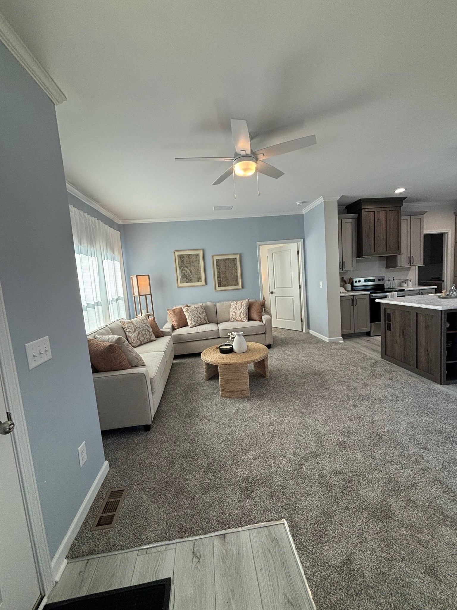 Cozy living room with a light gray L-shaped sofa and beige cushions. A round wooden coffee table sits on plush carpet. Soft blue walls and gentle lighting create a calm atmosphere.