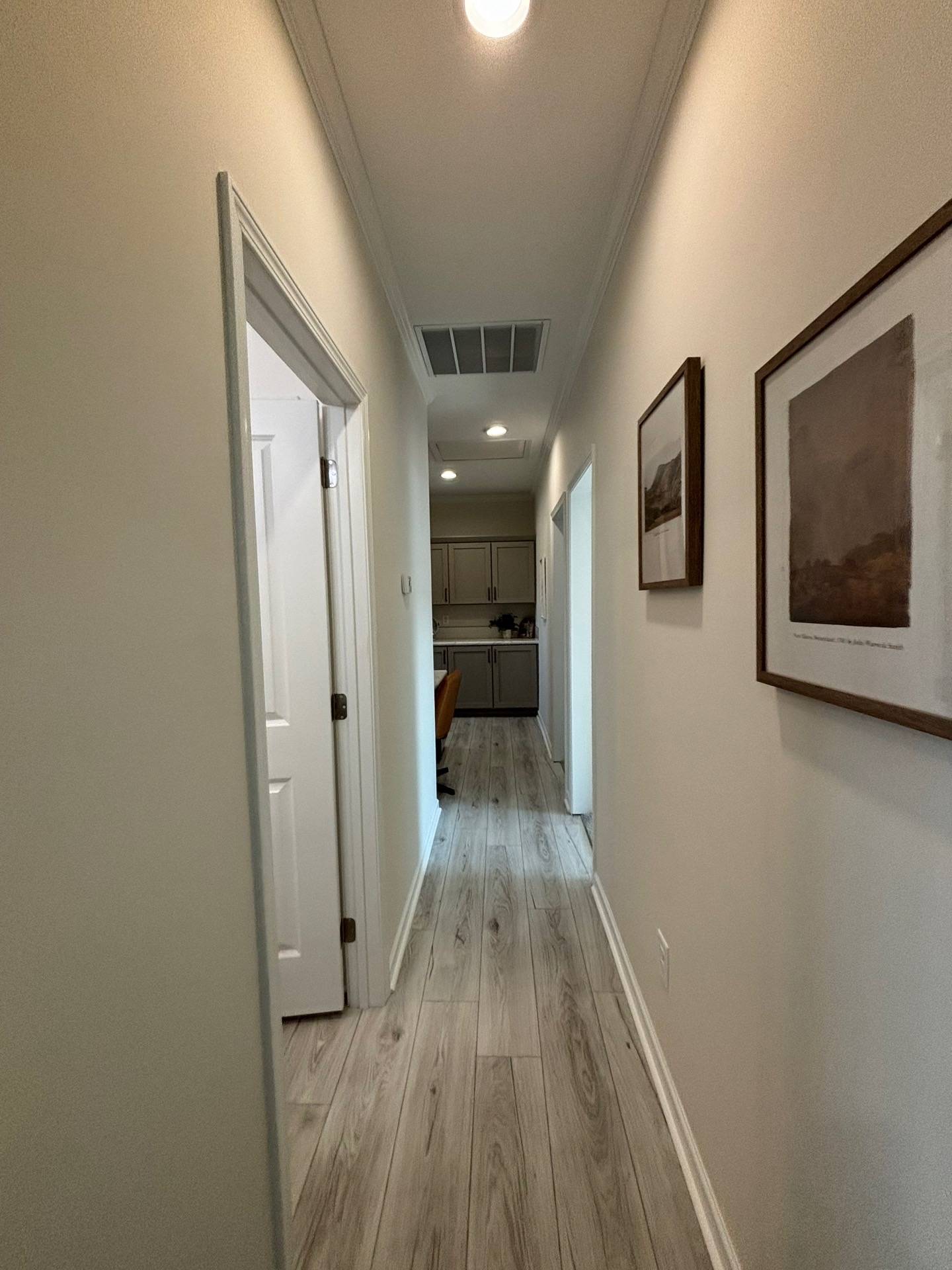 Long hallway with light wood flooring, white walls, and framed art. View leads to a room with kitchen cabinets. Bright, spacious, and orderly atmosphere.