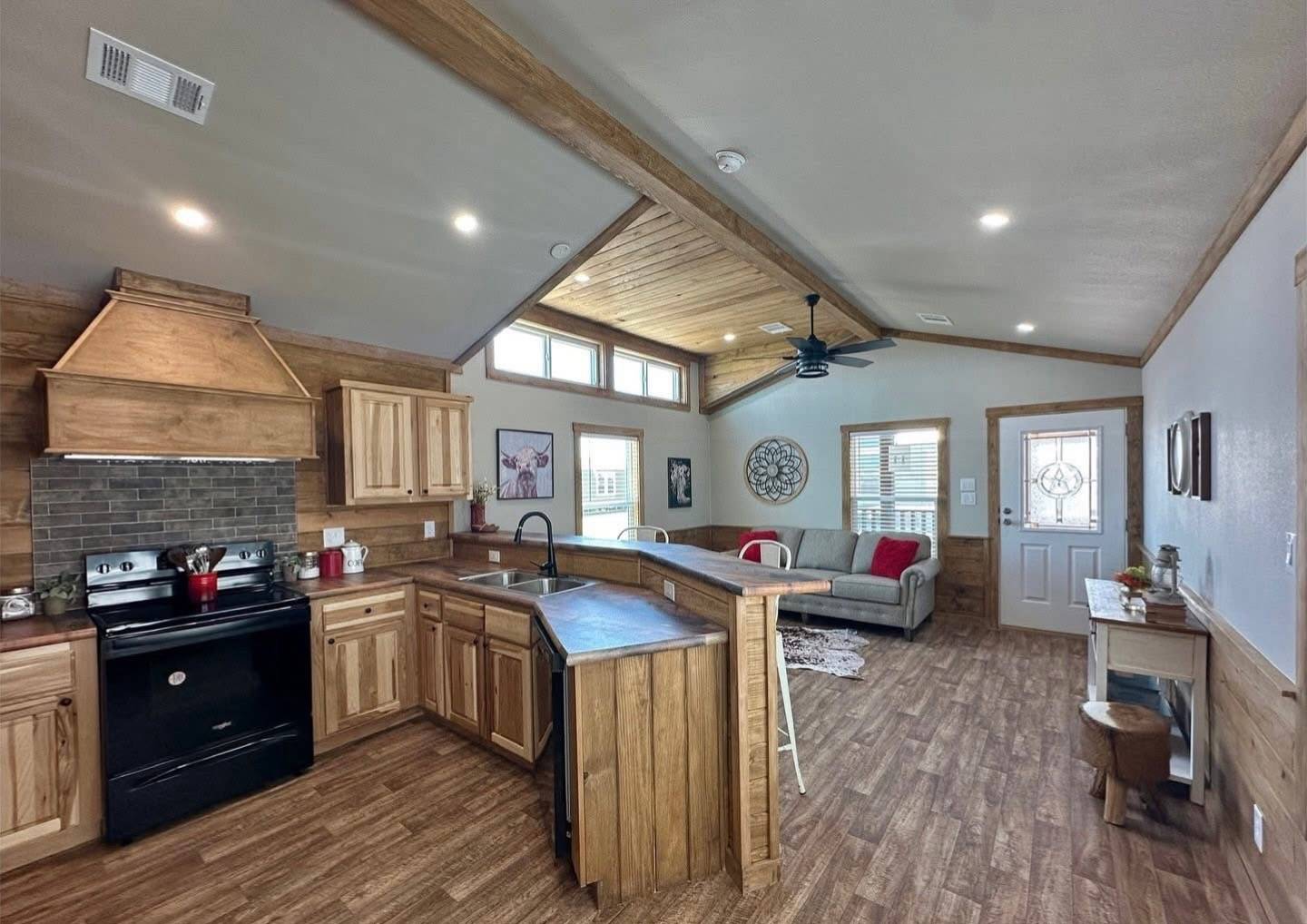 Spacious open-concept kitchen and living room with rustic wooden cabinets, dark appliances, sectional sofa, decorative wall art, and natural light.