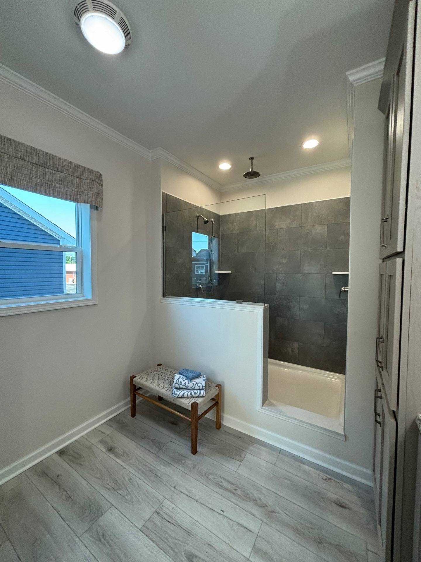 Modern bathroom with gray tiled shower, bench with folded towels, and light wood flooring. A window adds natural light, creating a serene atmosphere.
