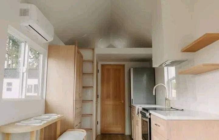 Compact modern kitchen with light wood cabinets, sleek countertops, and stainless steel appliances. Sunlight streams through a window, creating a warm, airy feel.