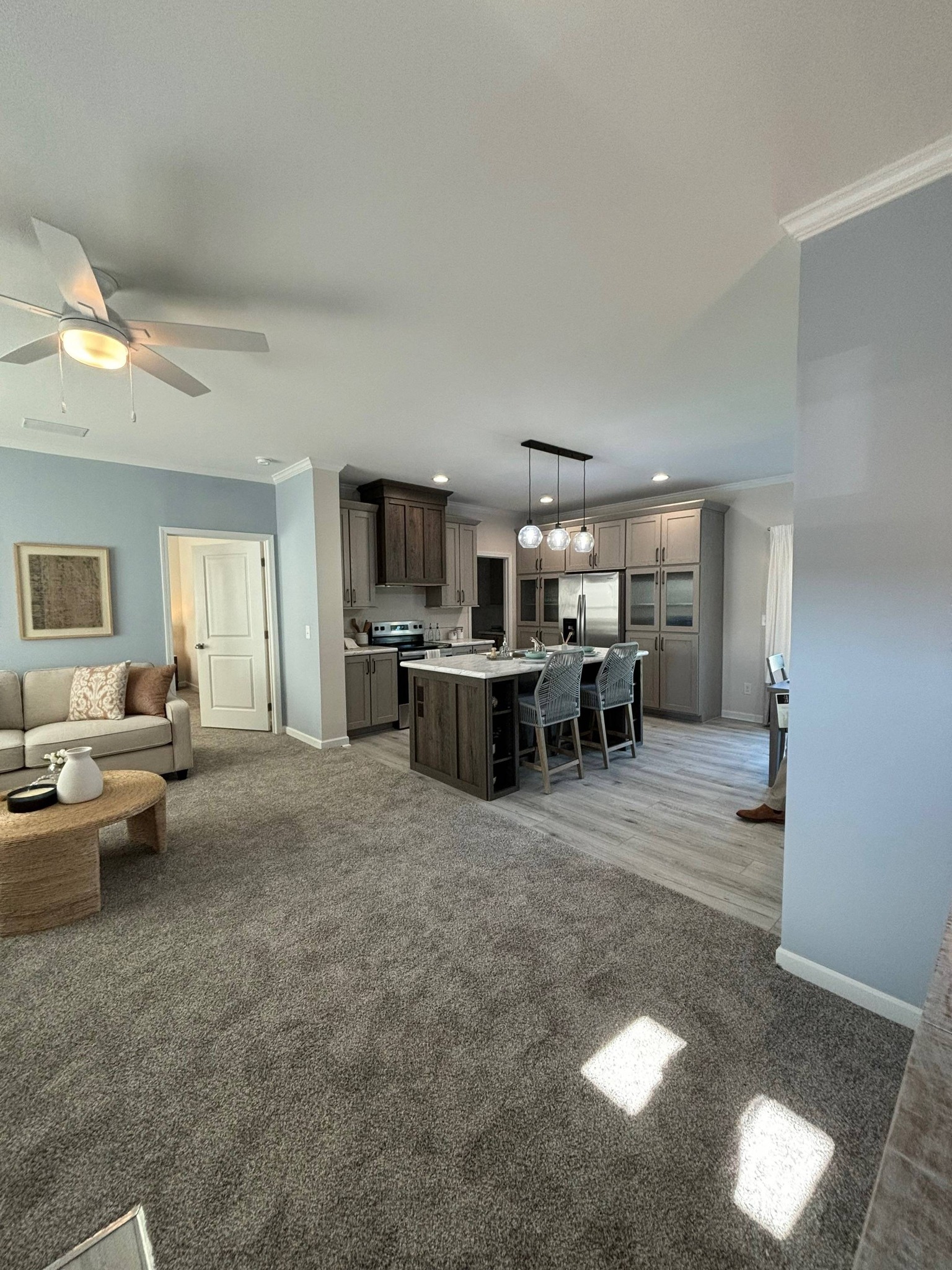 Open-concept living room and kitchen with cozy gray carpet, modern gray cabinets, island with bar stools, sofa, ceiling fan, and pendant lights. Calm and inviting ambiance.