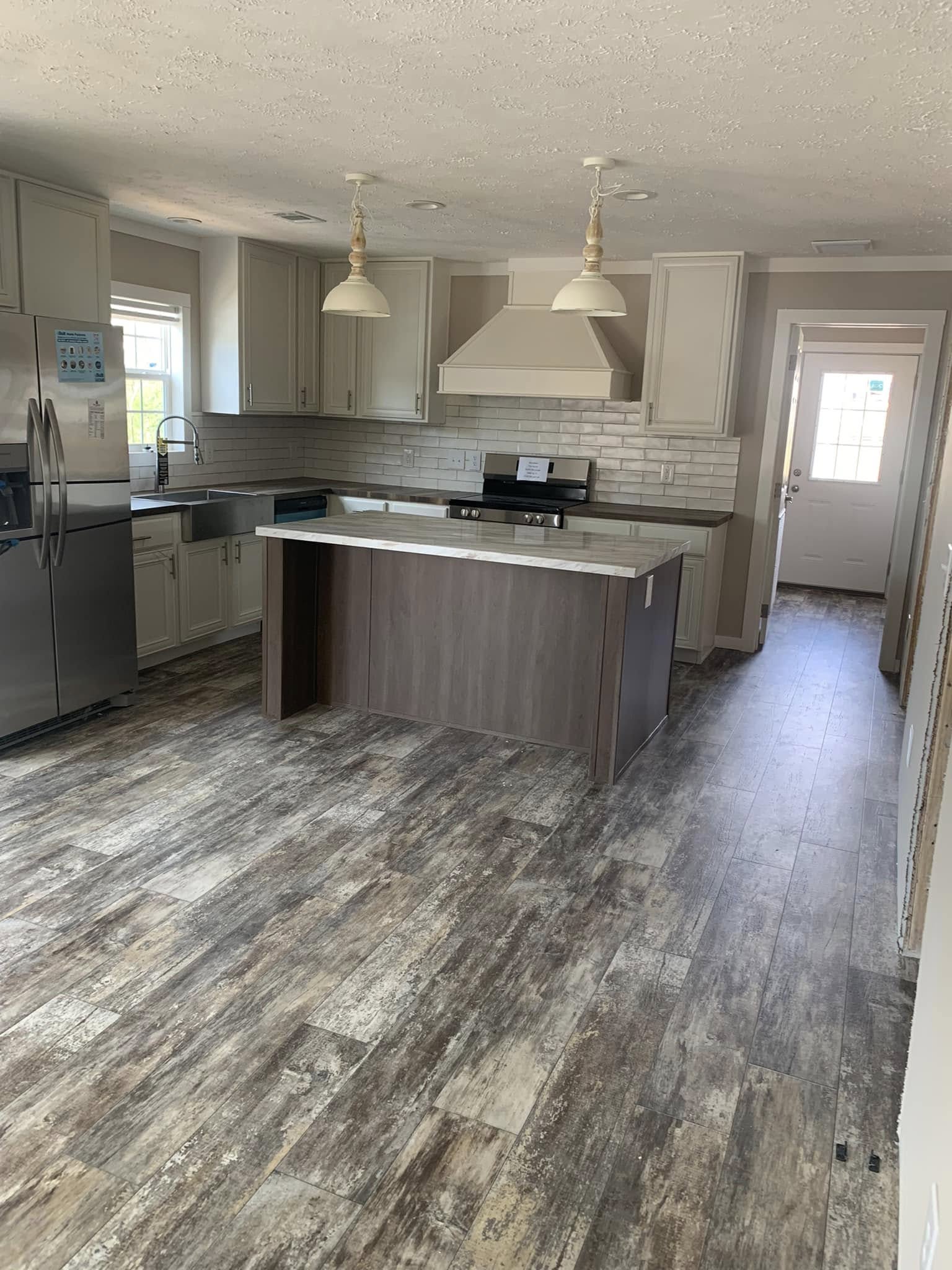 Modern kitchen with wood-look flooring, light cabinets, and stainless-steel appliances. Central island with pendant lights, door leading outside.