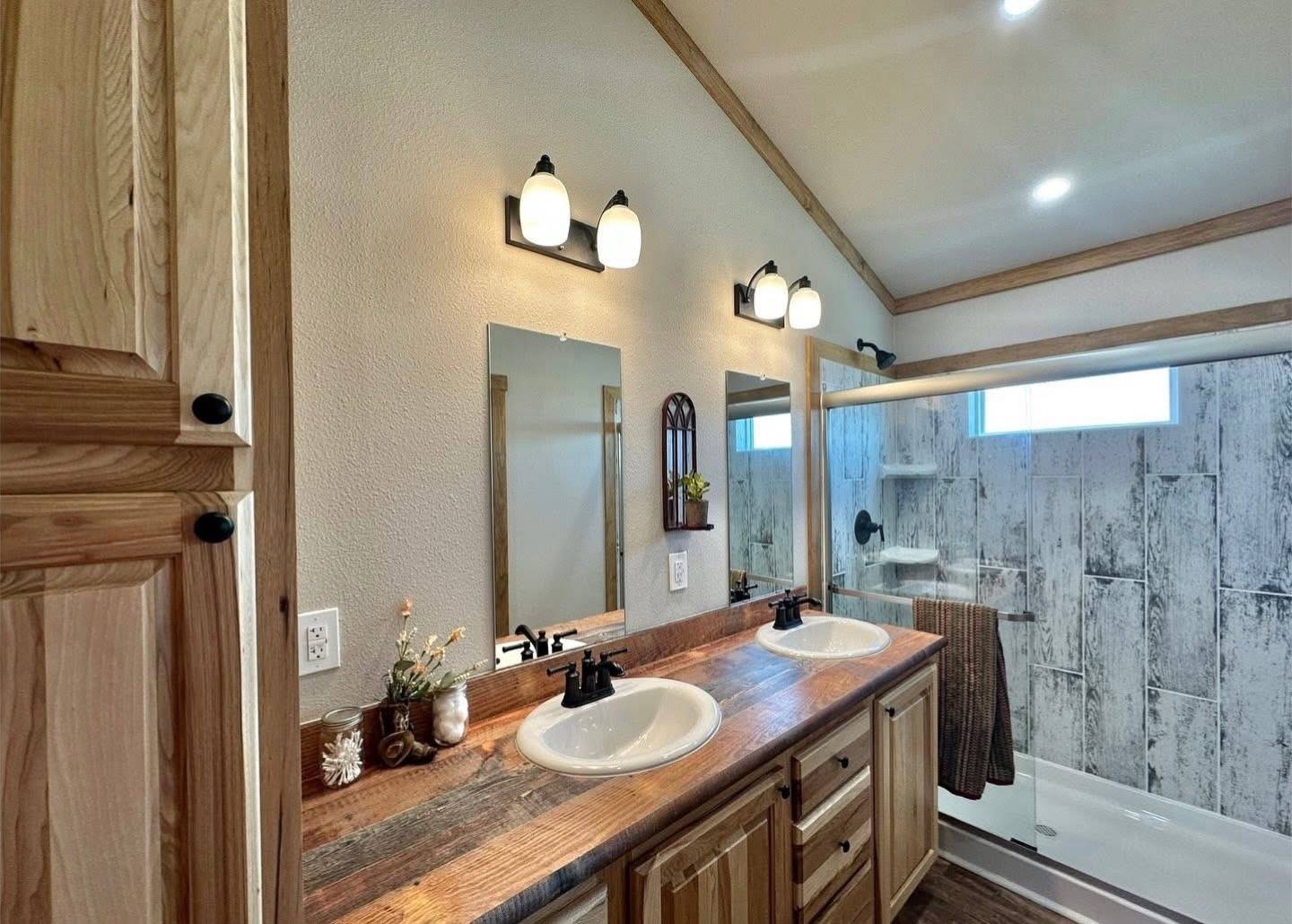 Rustic bathroom with wooden cabinets and countertops, dual sinks, and three warm lights above the mirrors. Shower area has a stone-like tile wall.