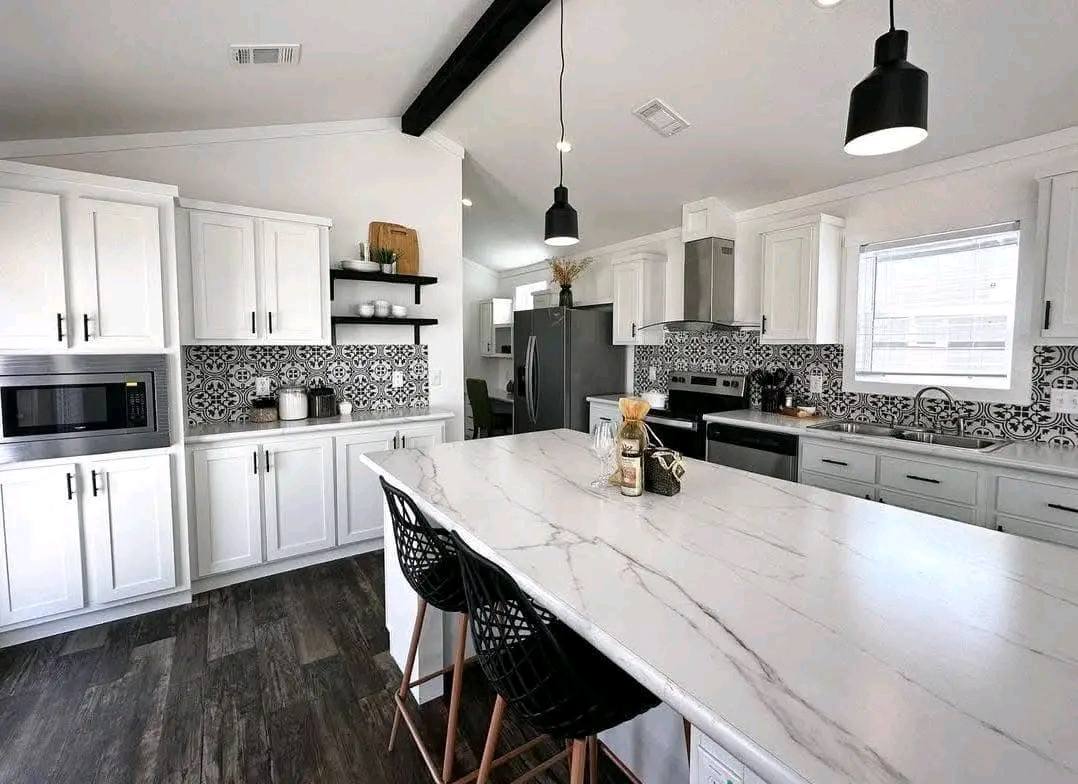 Modern kitchen with white cabinets, black-and-white patterned backsplash, dark wood floors, and pendant lights. Marble island with wicker chairs.